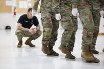 Partially uniformed man squats behind a group of uniformed soldiers inspecting their marching steps