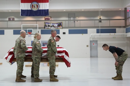 Six uniformed Pallbearers hold a flag covered coffin as another partially uniformed man inspects
