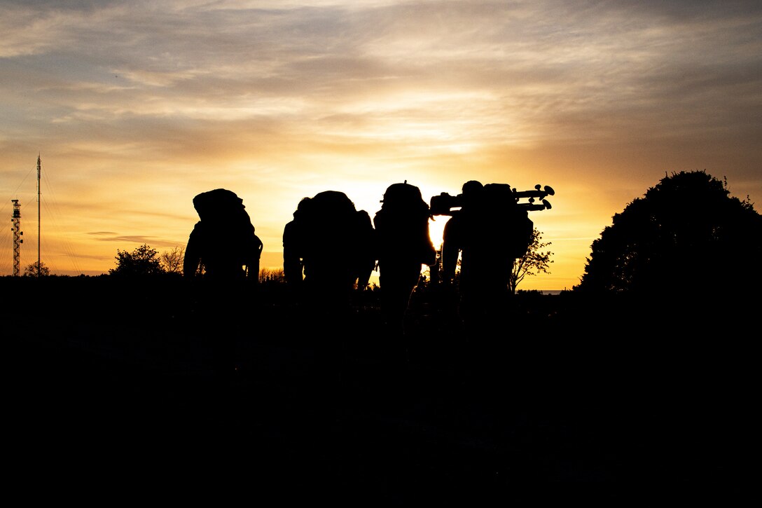 Several soldiers carrying gear across their backs are seen in silhouette at sunset.