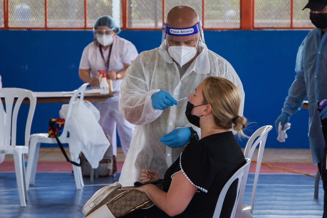 A soldier wearing personal protective equipment holds a swab to the nose of a woman seated beside him.