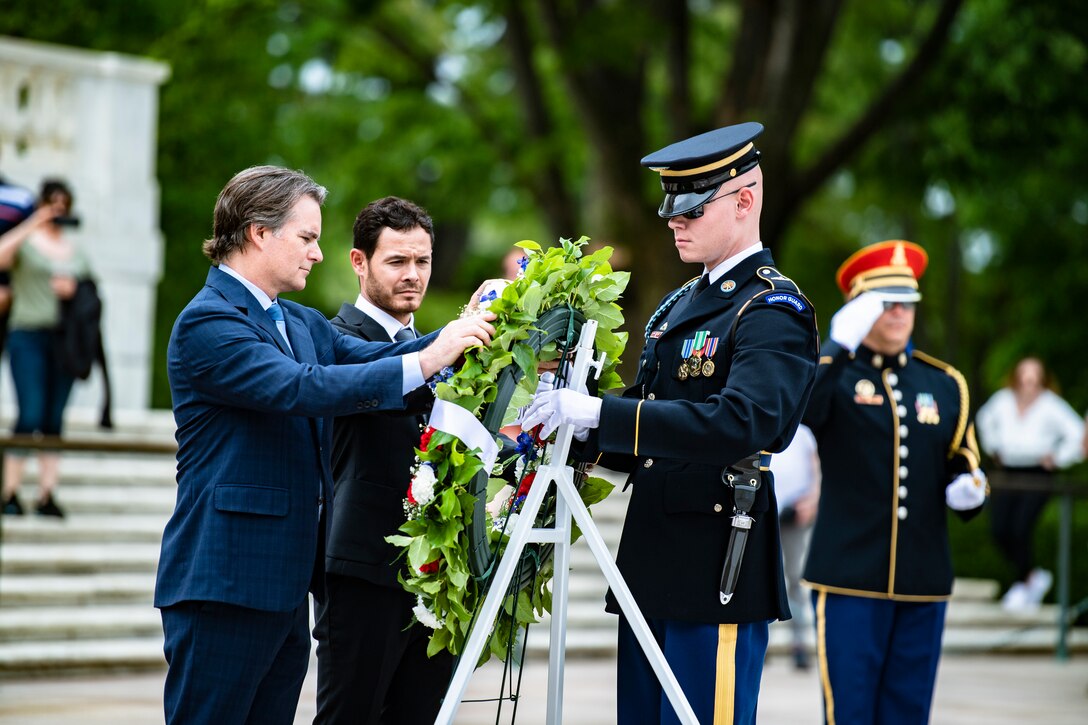 Two men in suits, and a man in a military uniform, adjust a wreath on a stand.