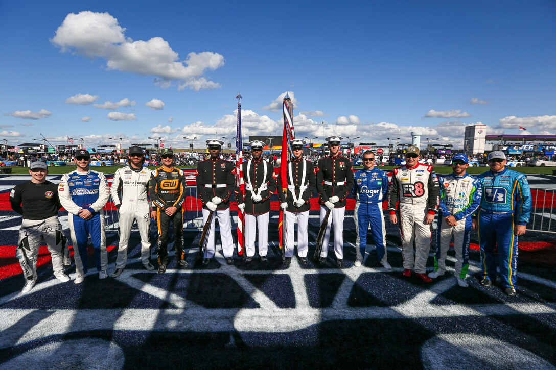 Service members in uniform and racecar drivers stand in a formation at a racetrack. One holds a flag.