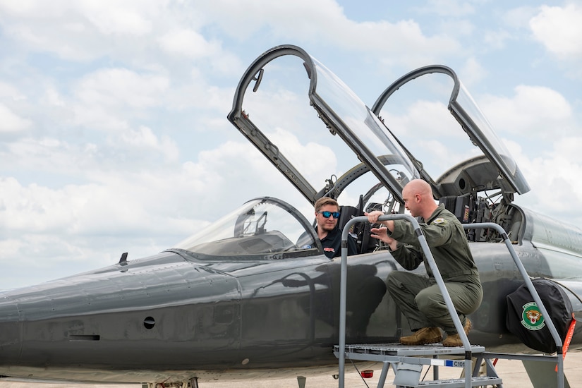 A man in civilian clothes sits in the cockpit of a fighter jet. A man in a uniform kneels on a ladder next to the aircraft.