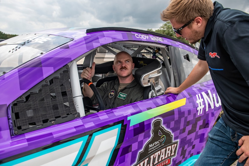 A service members sits inside a purple race car. A man stands near the driver’s side door.