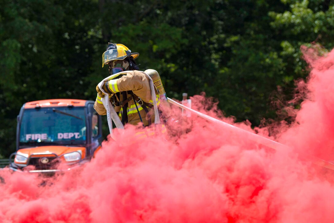 An airman wearing fire protective gear holds a hose as pink smoke surrounds.