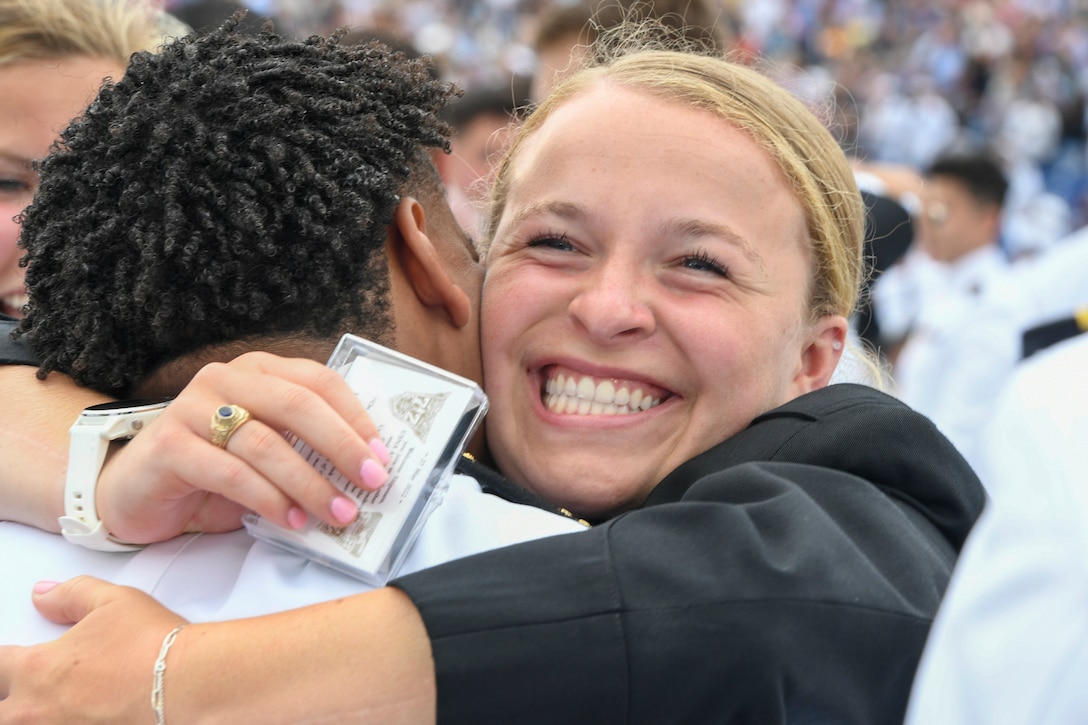 Two U.S. Naval Academy graduates hug while surrounded by others.