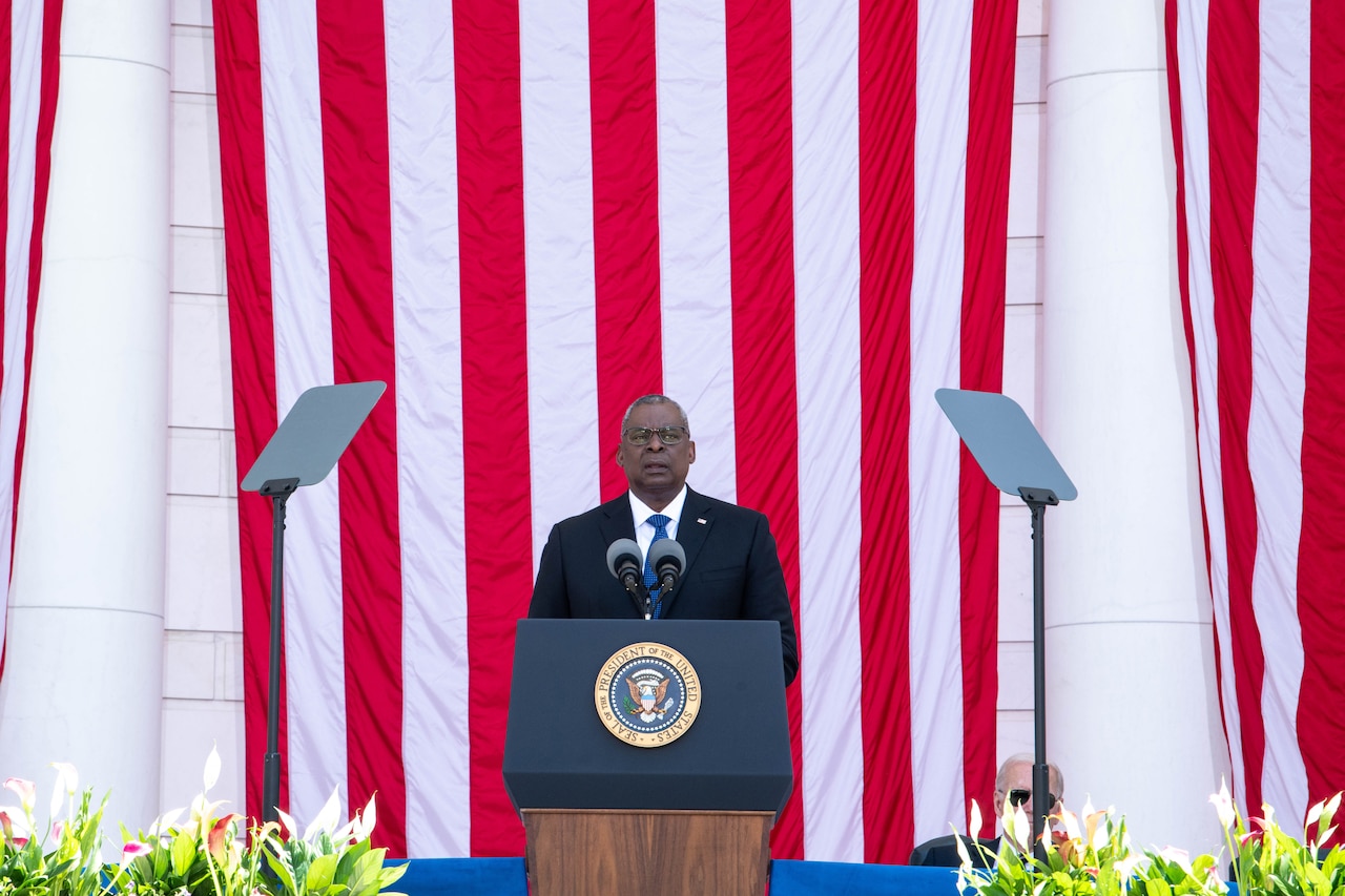 Secretary of Defense Lloyd J. Austin III speaks from a stage with large American flags behind him.