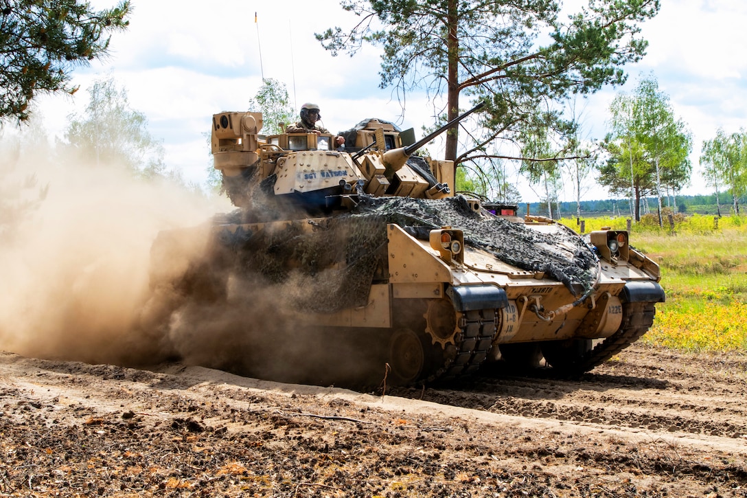A uniformed service member, sitting in the top of a tank, rides down a dirt path in a tank.