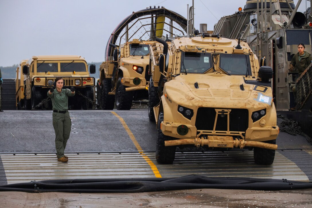 A service member guides a military vehicle down a ramp while another service member looks on.