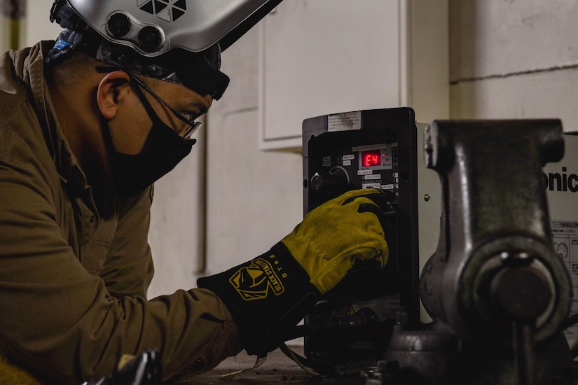 A sailor in protective welding gear manually adjusts settings on a device.