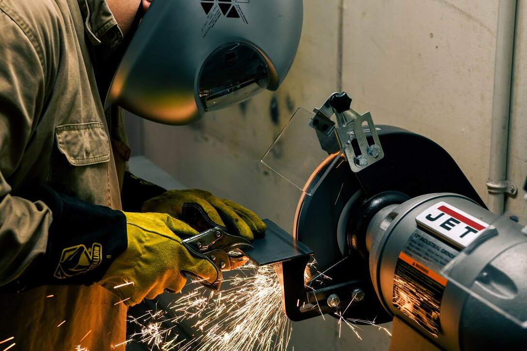 A sailor wearing a helmet grinds metal at a machine, creating sparks.