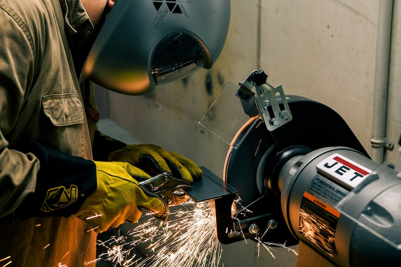A sailor wearing a helmet grinds metal at a machine, creating sparks.