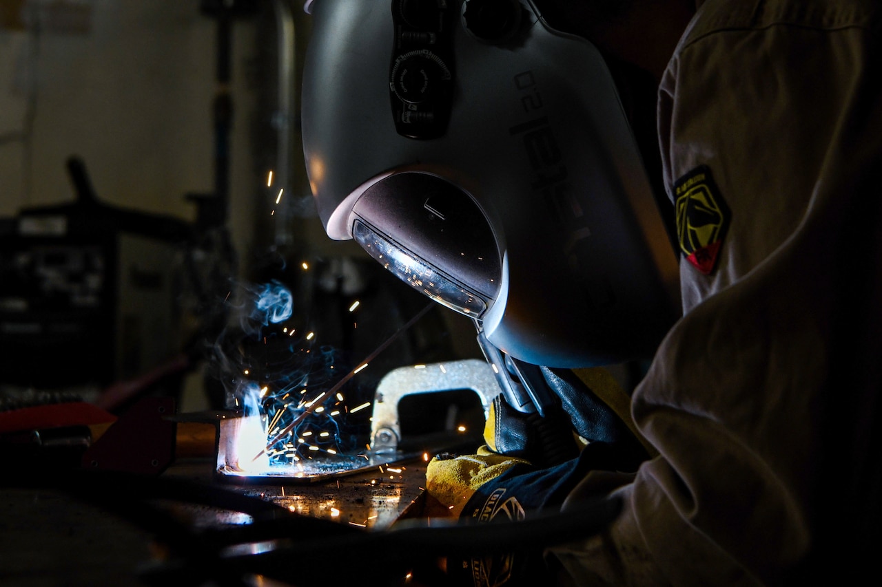 A sailor shown in profile in a protective helmet leans over his work as he welds.