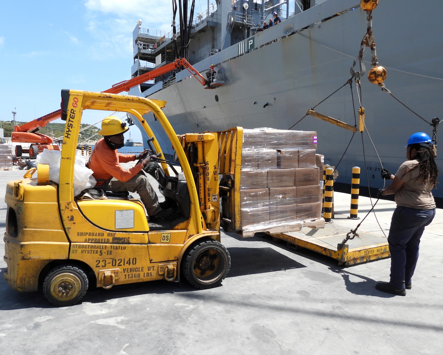 A man drives a forklift that is lifting a pallet of boxes while a woman steadies a platform so that the cargo can be lifted by crane onto the USNS Supply.