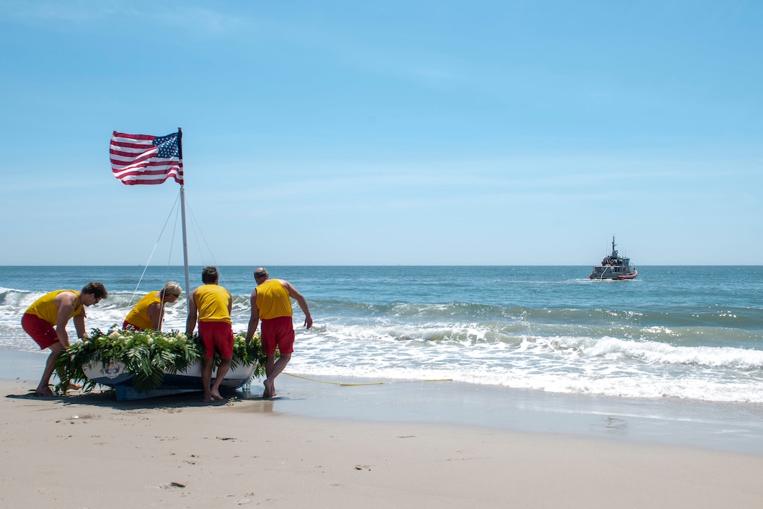 Four men carry a small boat covered with flowers and displaying an American flag into a body of water.