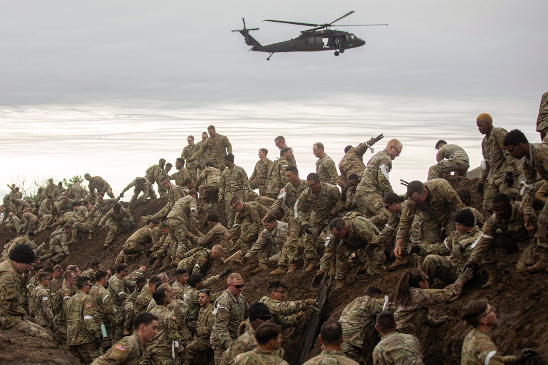 Soldiers climb out of a ditch as a helicopter flies above during an event.