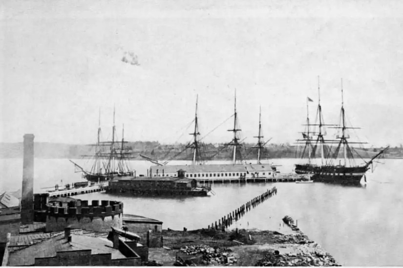 Four ships, three with masts, are moored at a dock.