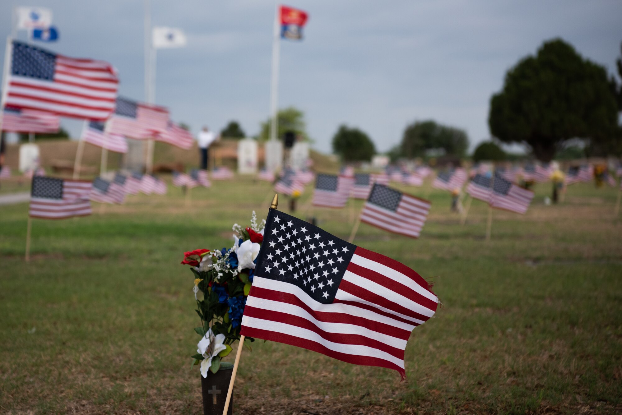 The American flag ruffles in the breeze alongside a vase of flowers atop a headstone at Lawnhaven Memorial Gardens, San Angelo, Texas, May 30, 2022. Flags were planted by volunteers from the San Angelo community and Lawnhaven staff at every service member’s grave in preparation of a Memorial Day ceremony (U.S. Air Force photo by Senior Airman Michael Bowman)