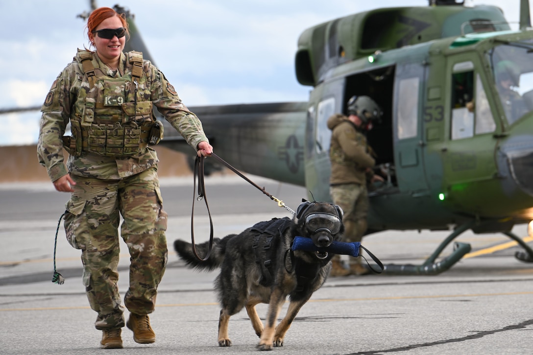 An airman walks with her dog on a leash with a helicopter behind them.