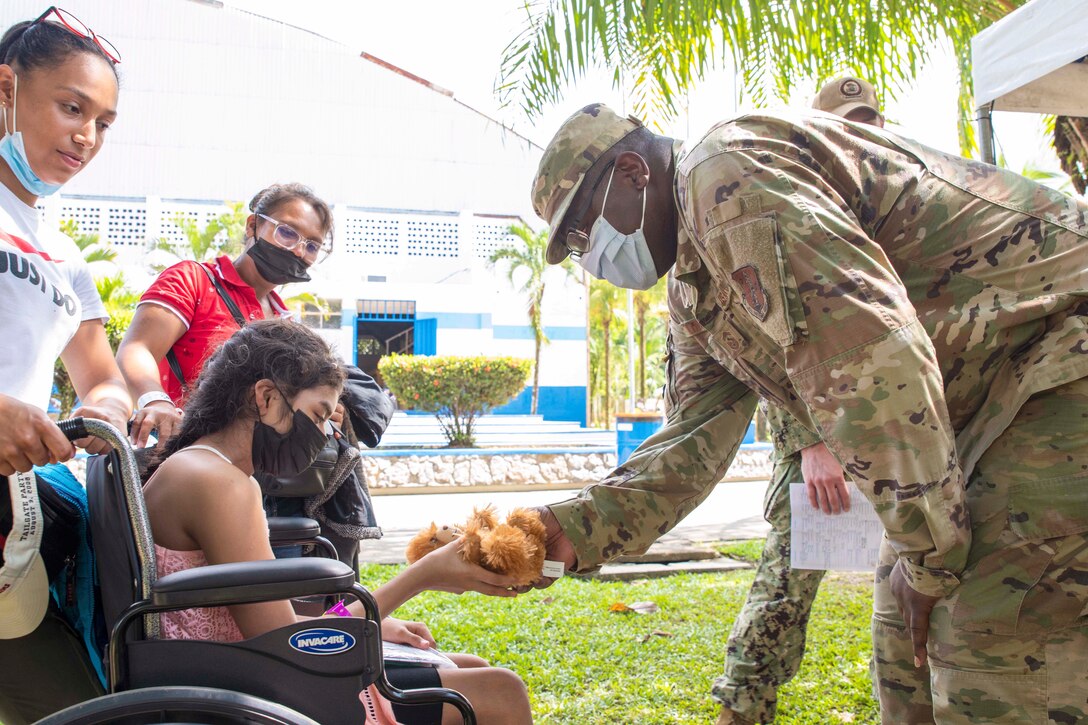 An airman gives a girl in a wheelchair a stuffed bear.