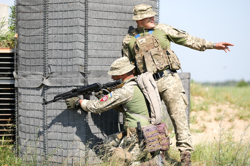 Two soldiers take a position behind cover. One is kneeling with a weapon pointed, and the other is looking back.