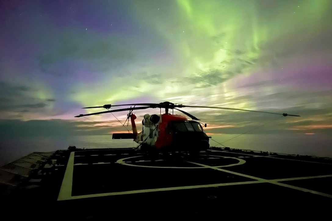 A helicopter sits on the deck of a ship with the northern lights in the backdrop.