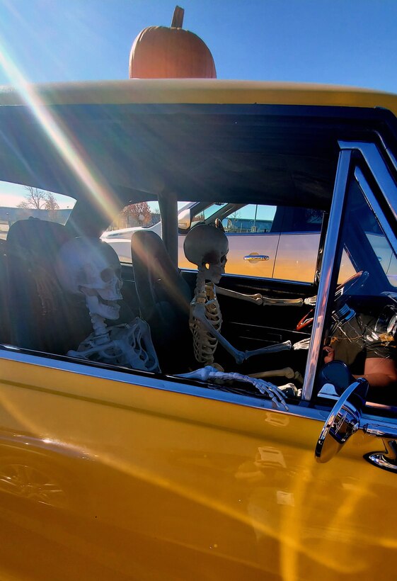 Two skeletons driving a car with a pumpkin on top. The car is yellow.