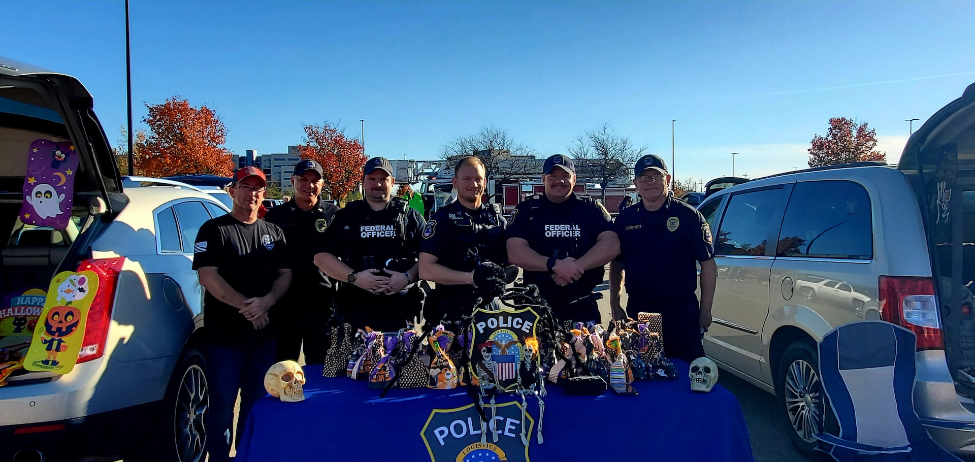 Several police officers in black police uniforms stand behind a table with a blue tablecloth and the DSCC/DLA Police logo and many treat bags displayed on top.