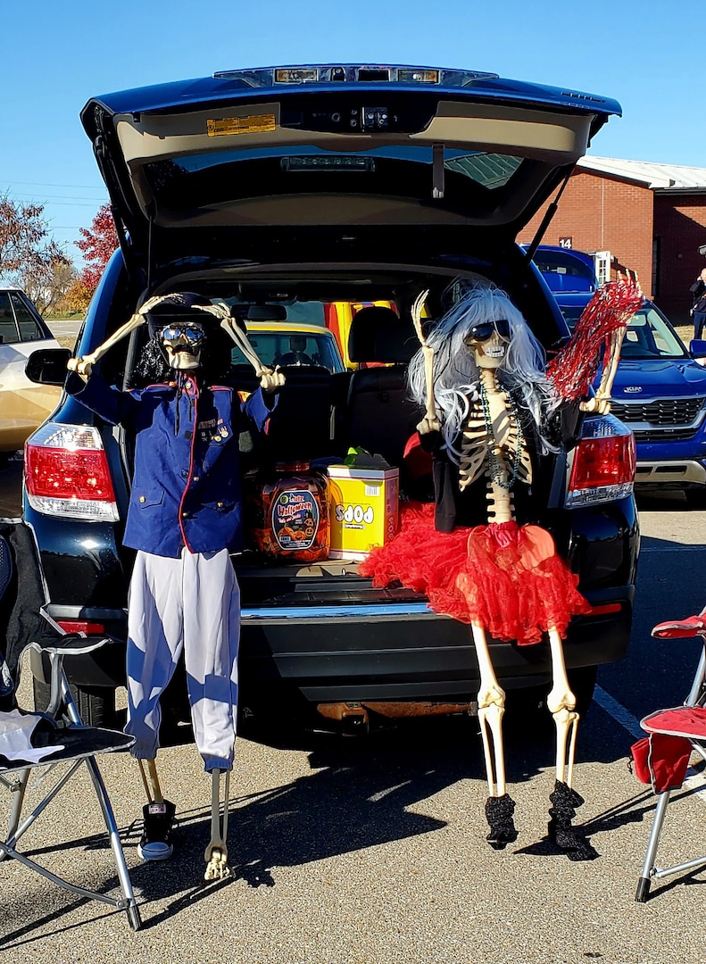 Two skeletons with costumes on flank a trunk filled with treats. One has a red dress on and the other a baseball outfit.