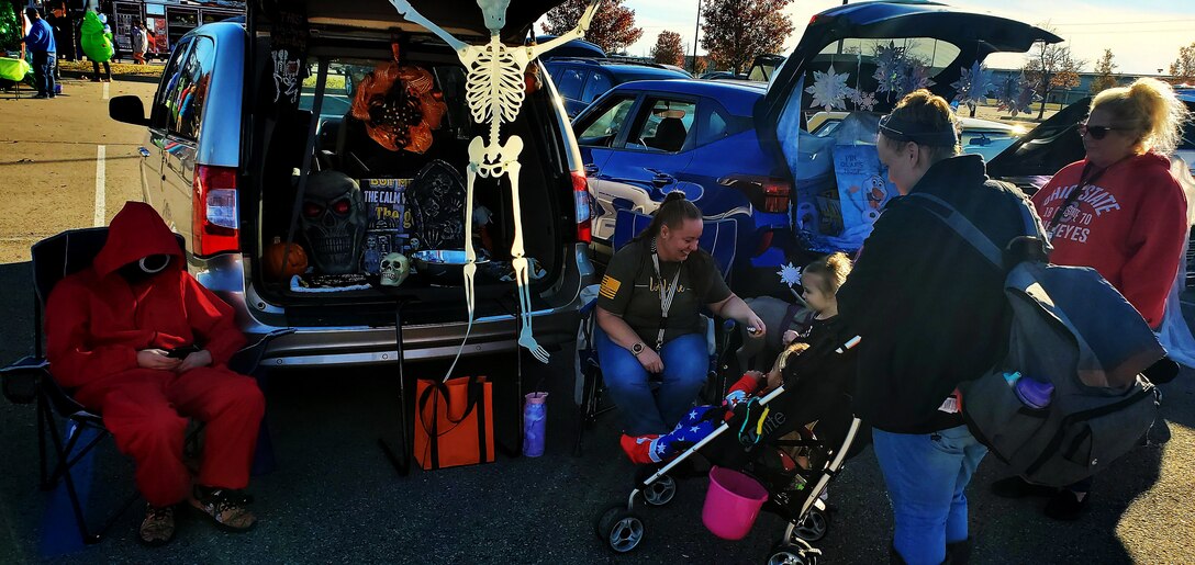 A person dressed in a red body suit on their phone sits next to a decorated trunk with treats while a lady with brown hair hands out treats on the other side to several kids and parents.