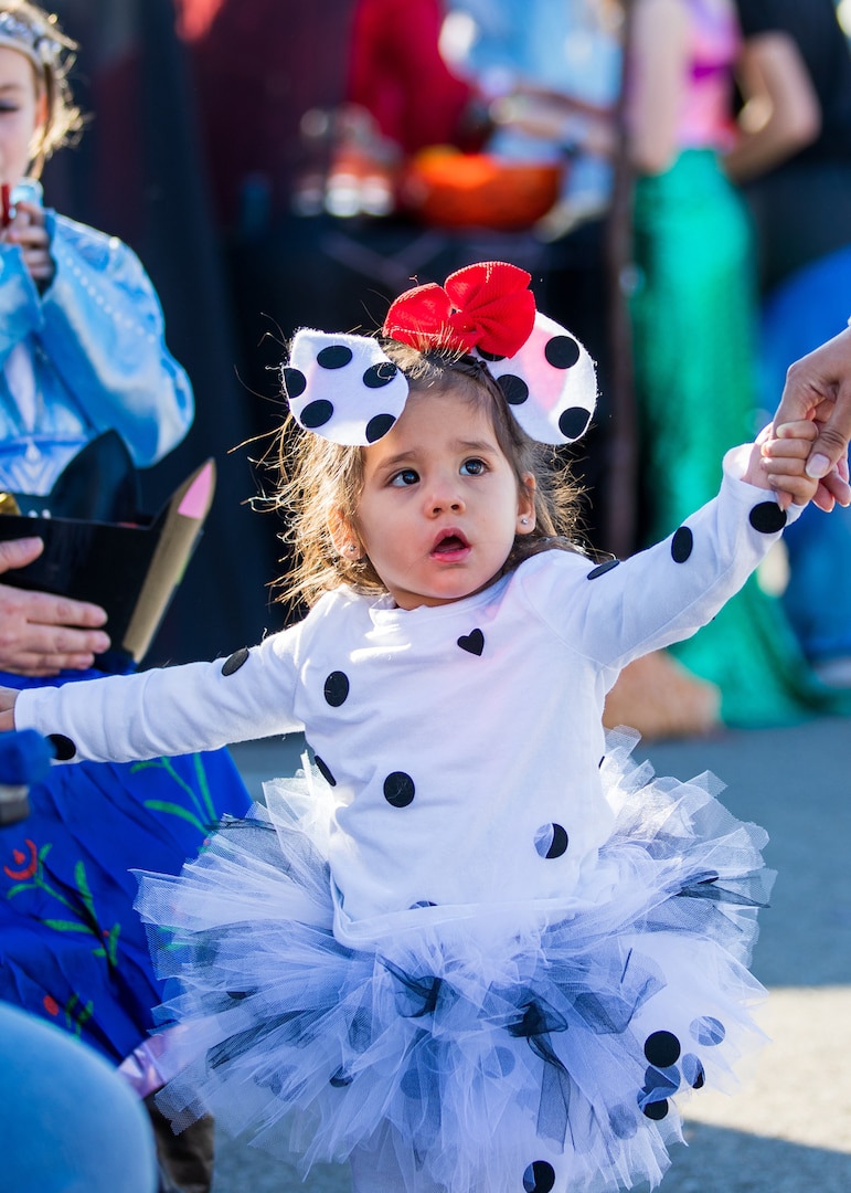 A little girl in a polka dot dress and tutu. It is mostly white with black polka dots. She has a bow on her head. She has brown hair and is being led by her parent.
