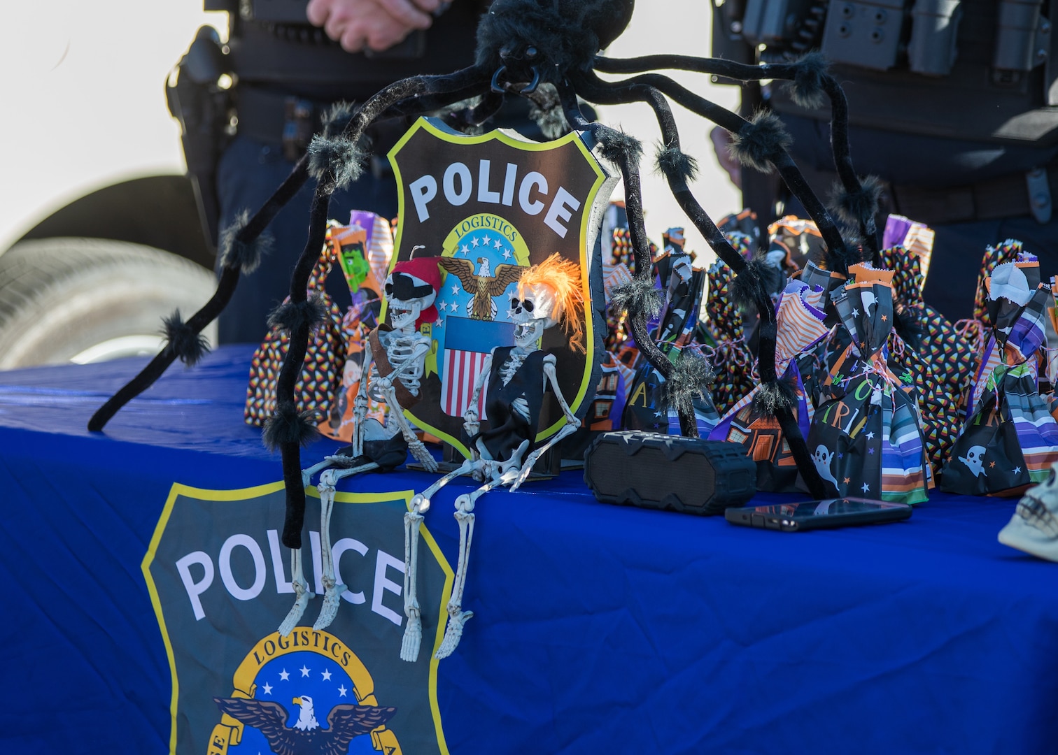 A table with a blue tablecloth with the DSCC/DLA Police logo on it with treat bags on top. The logo is flanked by mini skeletons.