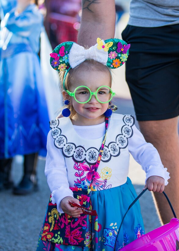 A girl with green rimmed glasses, a bow in her hair with butterflies and flowers all over her white dress.