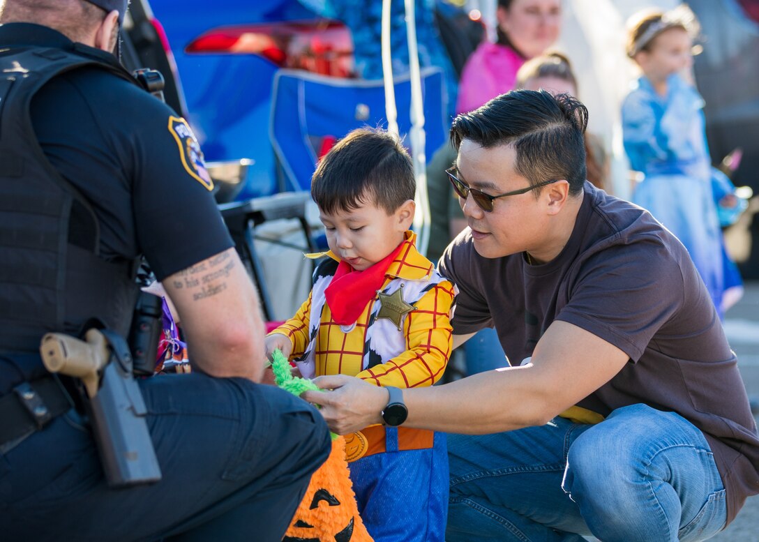 A boy dressed as Sherriff Woody from A Toy Story movie is getting a treat with the help of his parent, a man with black hair sunglasses and wearing a T-Shirt and blue jeans.