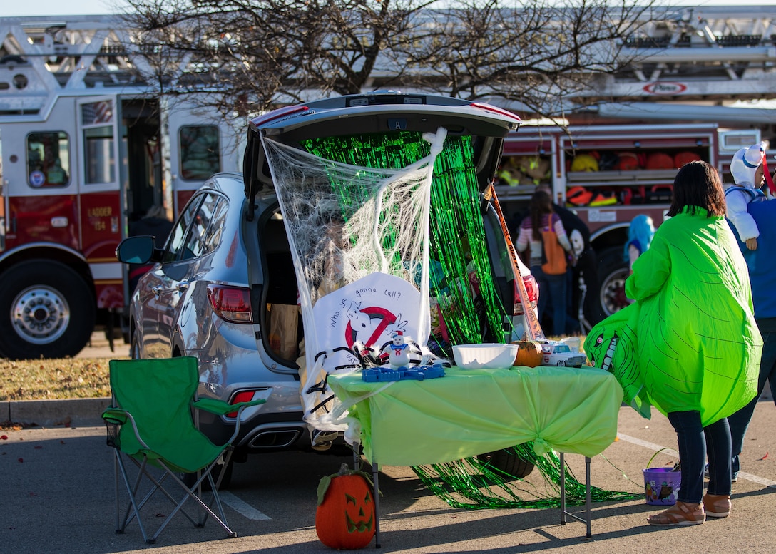 A trunk of a car with Ghostbusters the movie themed decorations. A woman dressed as the green goblin ghost stands next to it. Another person hides in the trunk behind a bunch of curtains of yarn and other fabric. A table sits outside with treats.