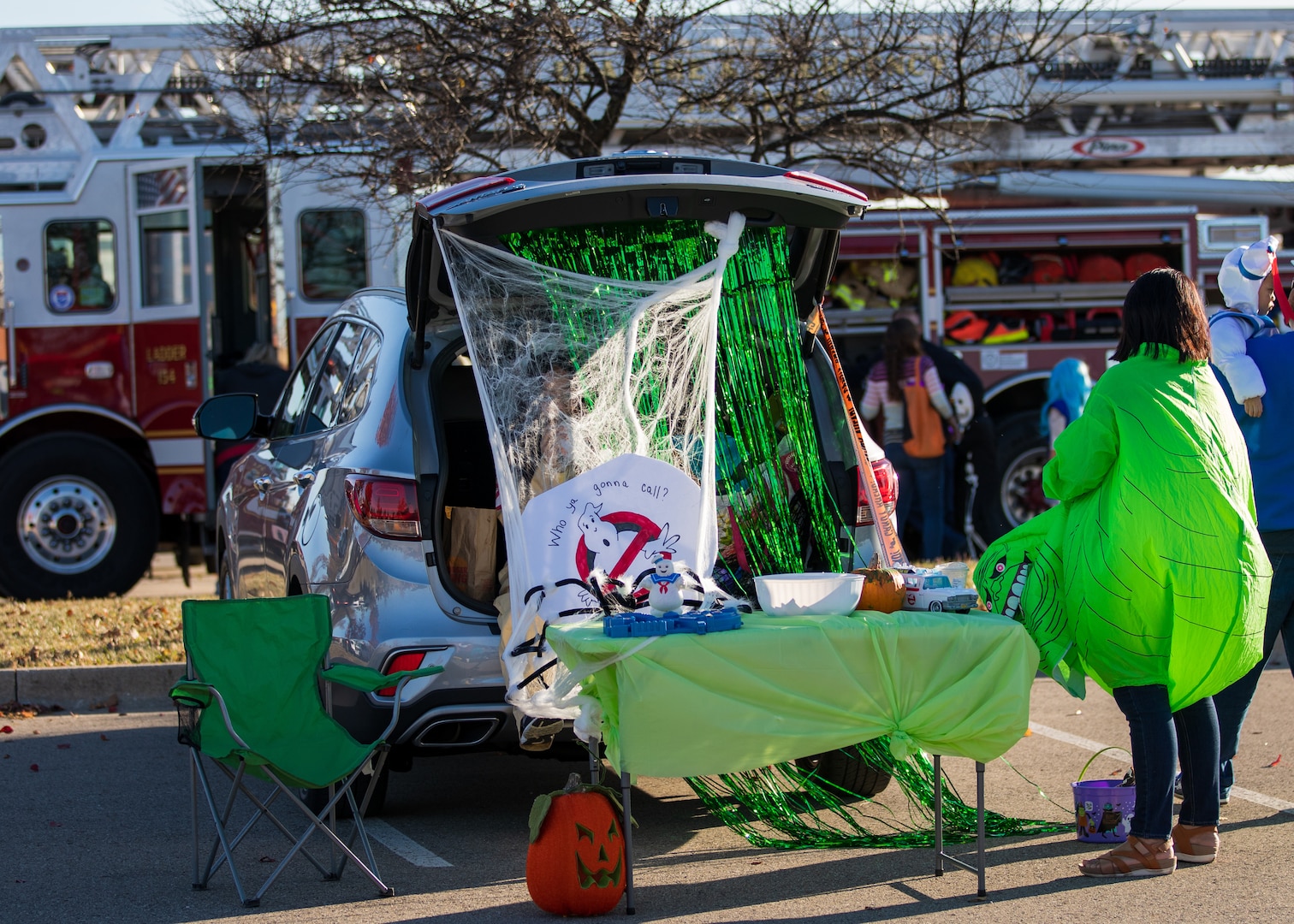 A trunk of a car with Ghostbusters the movie themed decorations. A woman dressed as the green goblin ghost stands next to it. Another person hides in the trunk behind a bunch of curtains of yarn and other fabric. A table sits outside with treats.