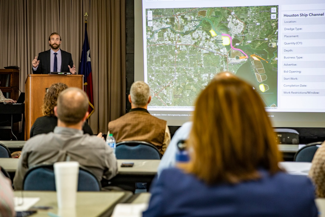 Man speaks at podium in front of audience next to a large video screen