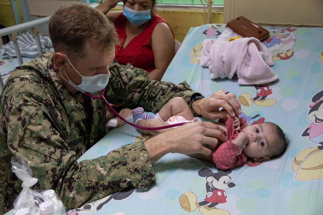 A military doctor checks the heart of a baby lying on a bed.