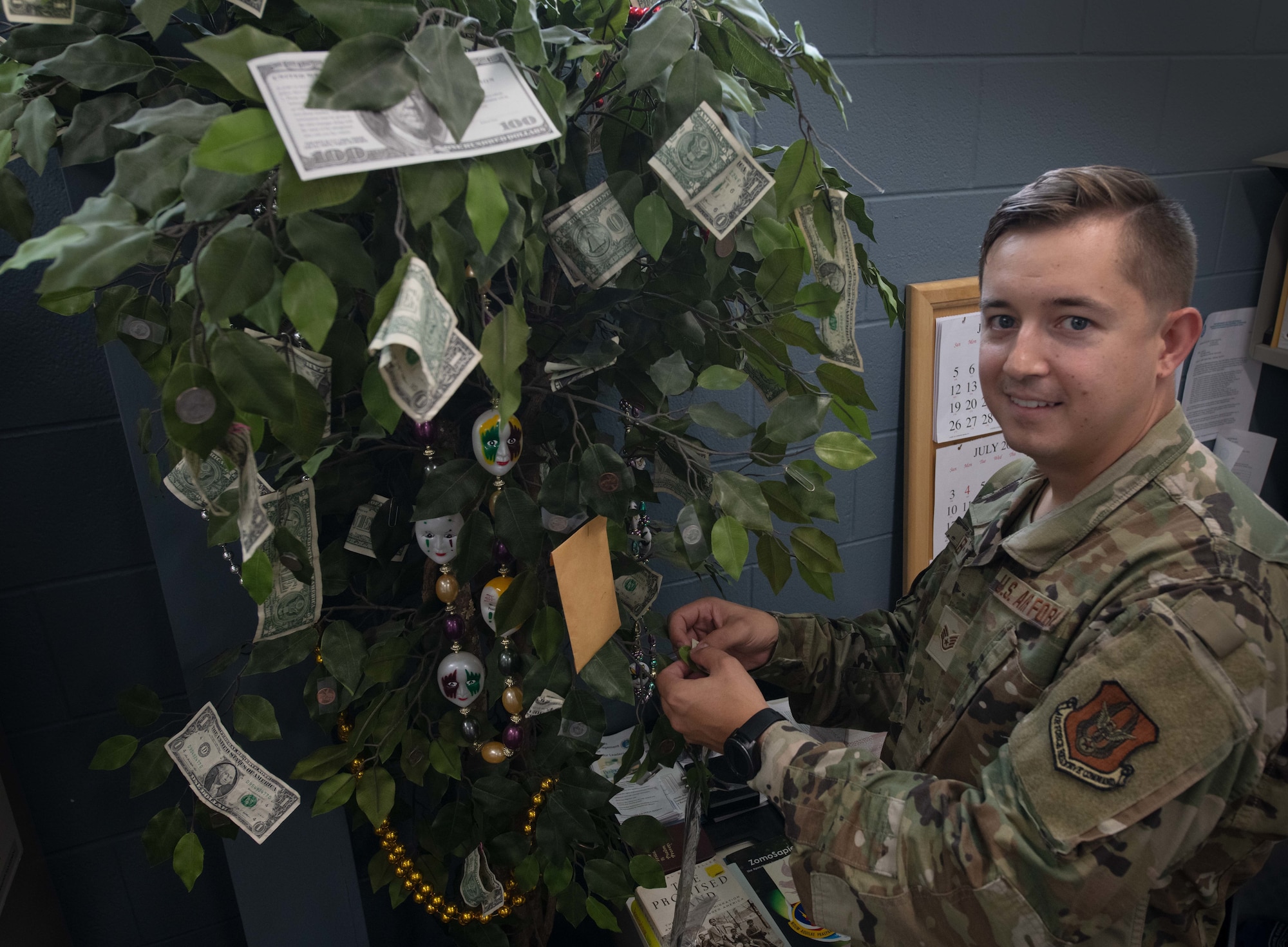An Airman attaches a dollar to an artificial tree, decorated with dollar bills inside an office.