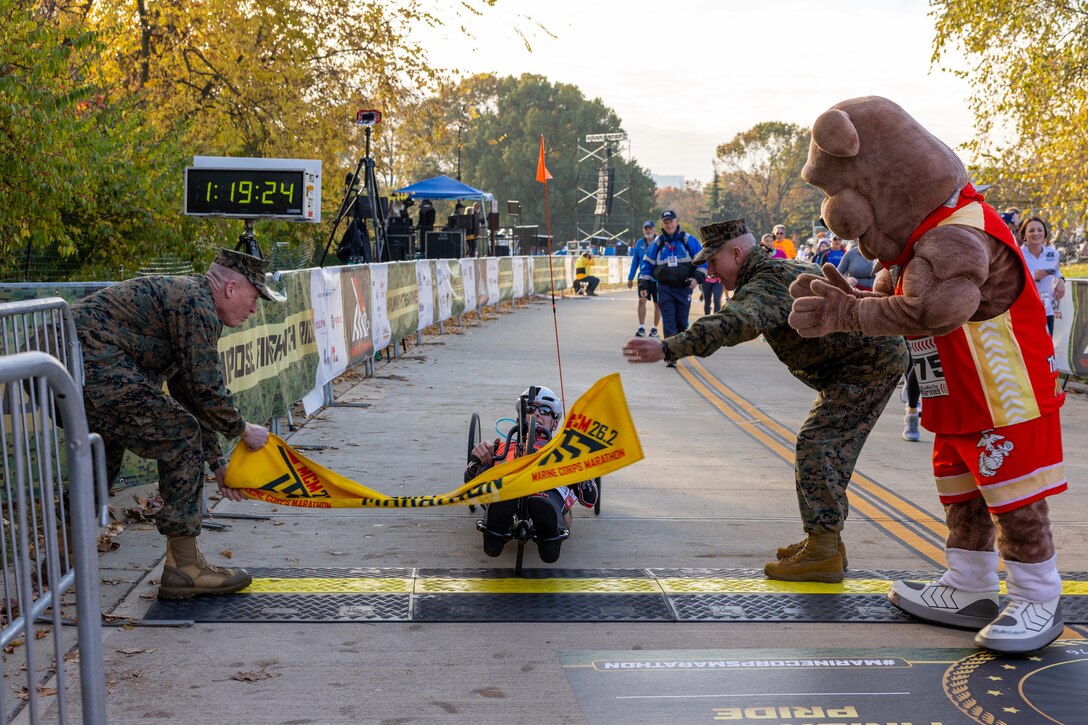 A hand cyclist pedals across the finish line in a race as two service members let go of a banner.
