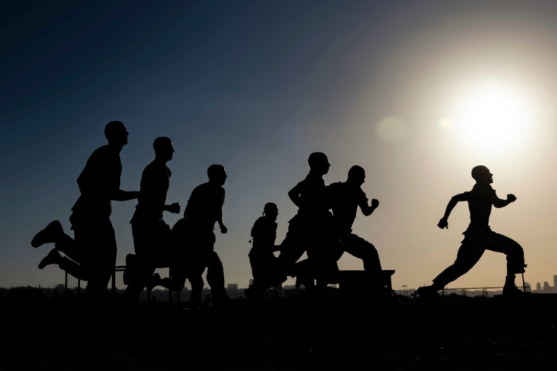 Marine Corps recruits run under a sunlit sky as shown in silhouette.