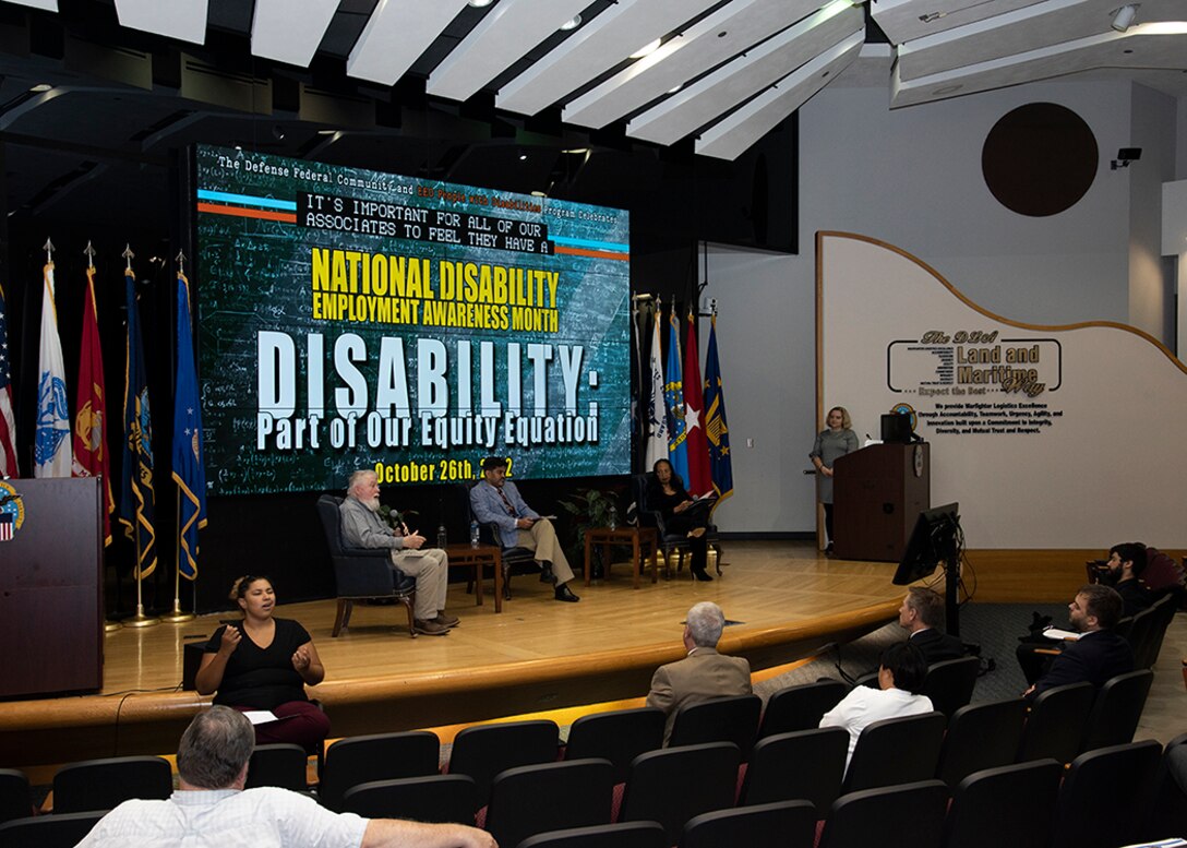 Two men and a woman are seated on stage while a second woman stands at a podium.