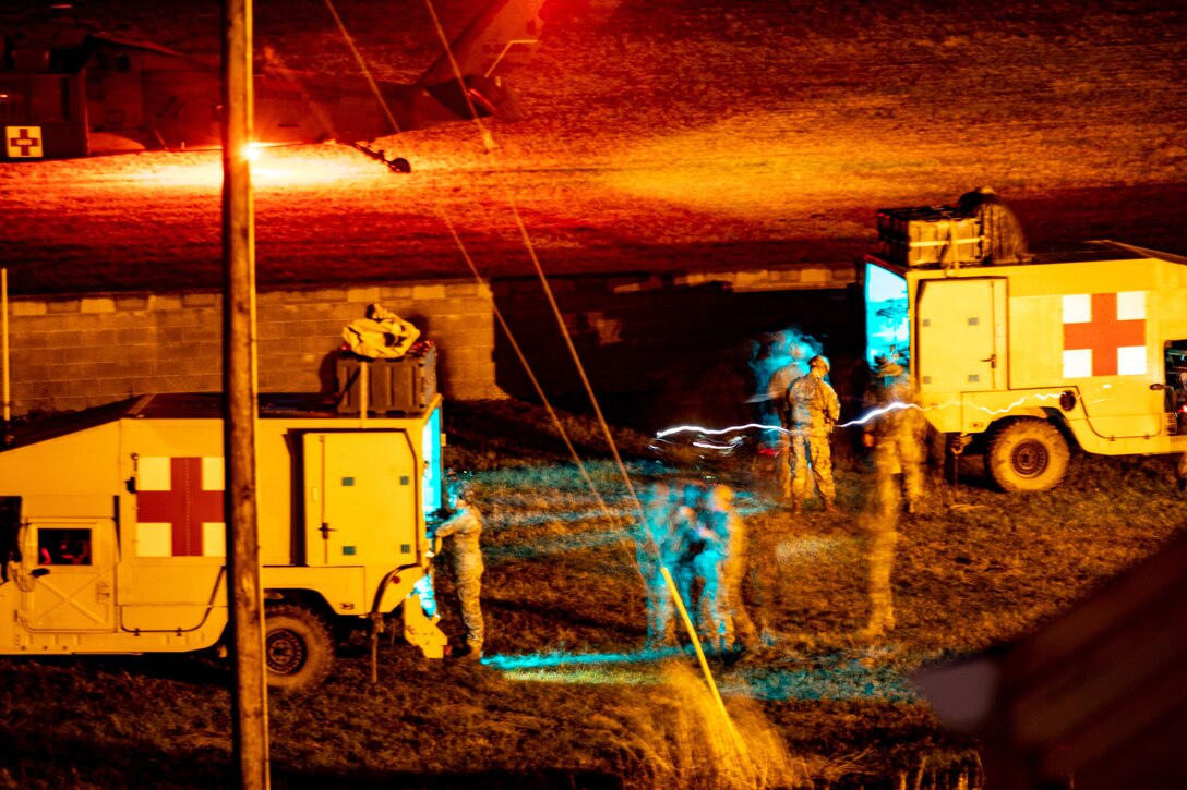 Two parked medical vehicles and service members stand in a brightly lit area.