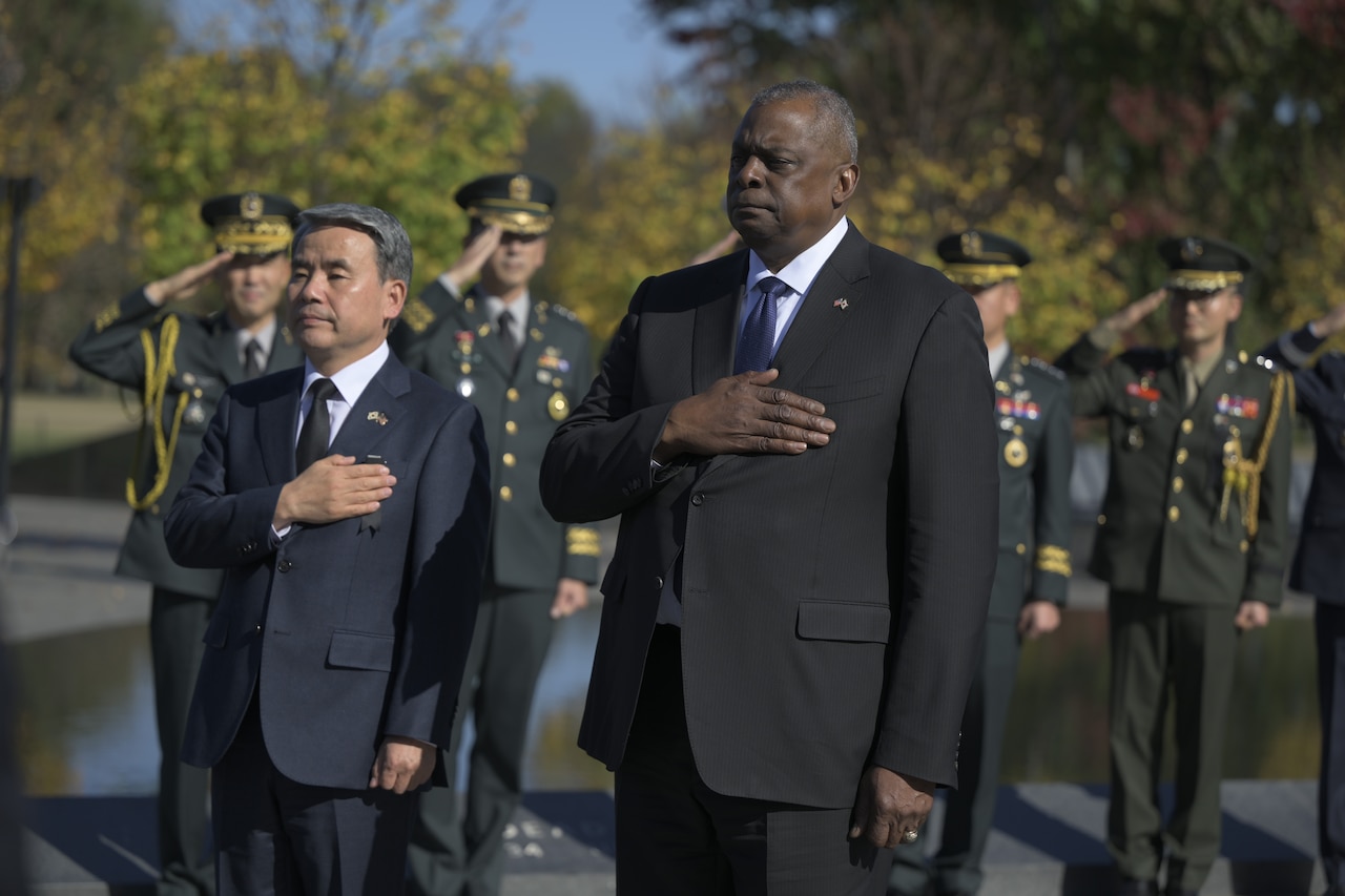 Two men hold their right hands over their hearts, as service members salute behind them.