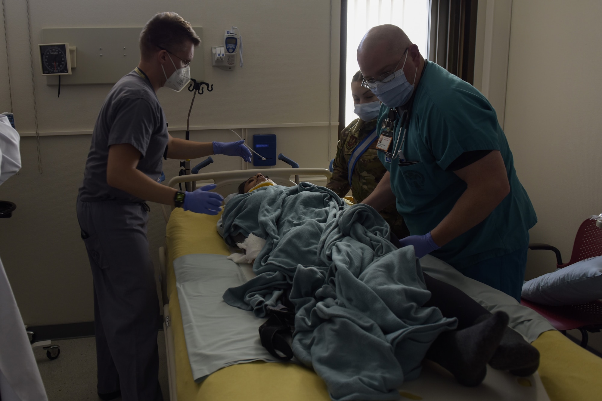 A woman acting injured lays on a hospital bed, while two men and one women tend to her injuries.