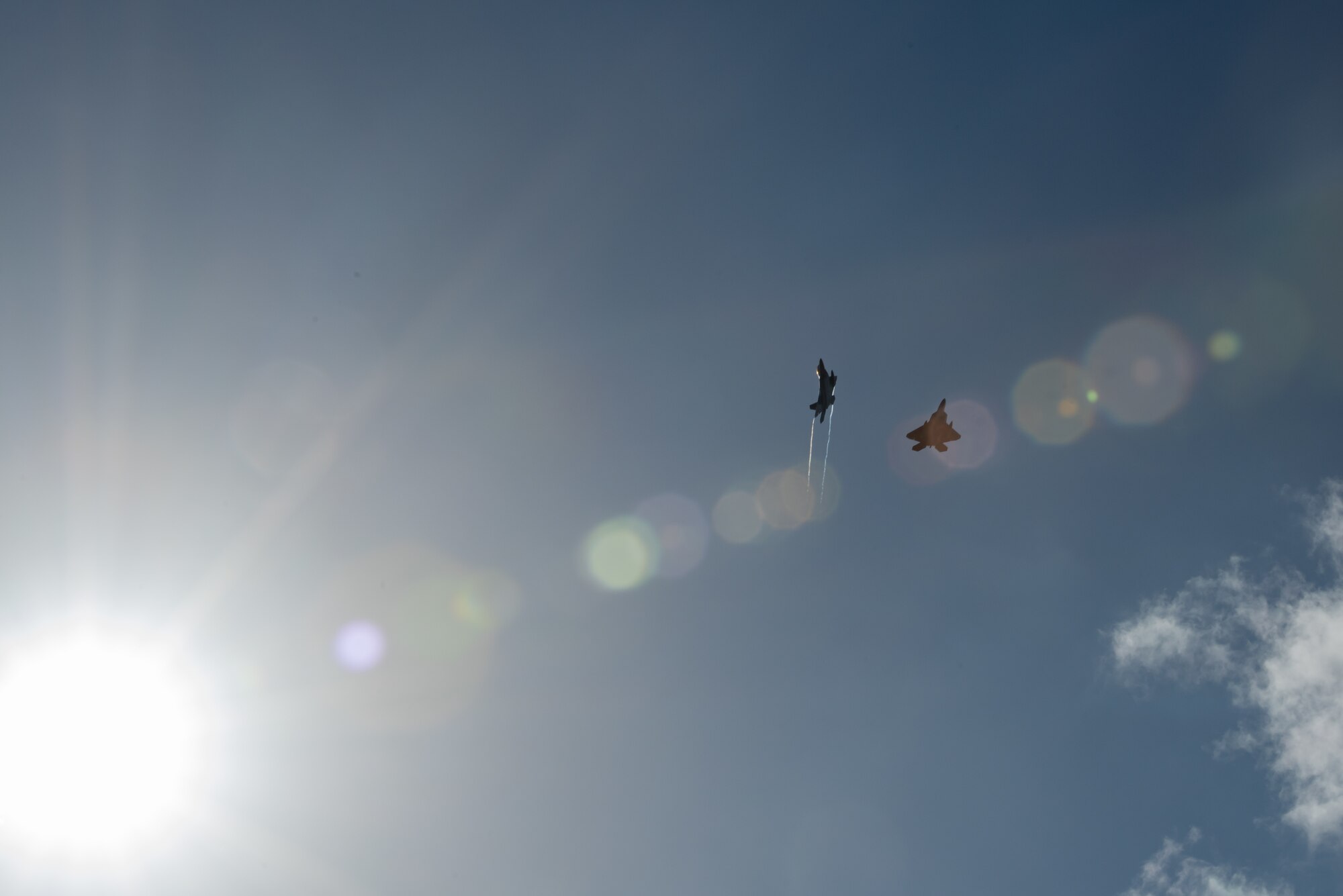 Two F-22 Raptors fly overhead with the sun in the lower left corner. One F-22 breaks away while the other F-22 flies straight ahead.