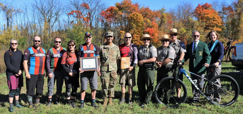 A group of individuals pose with a certificate/plaque in front of trees in a park-like setting