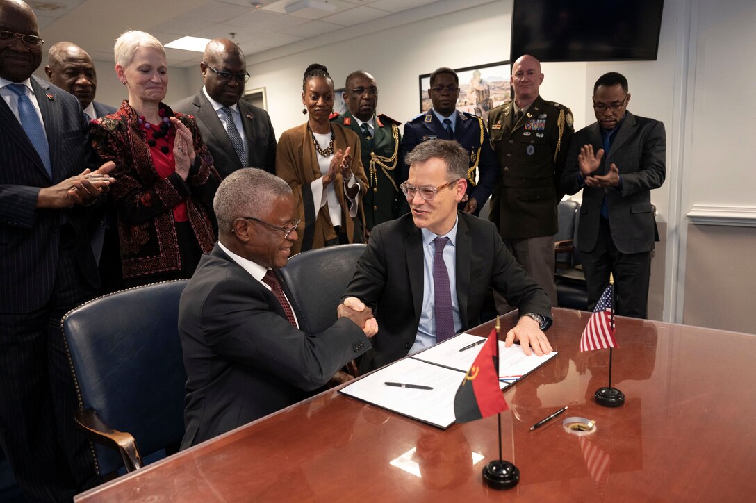 Two men sit next to one another at a table and shake hands; in front of them, are documents and pens.