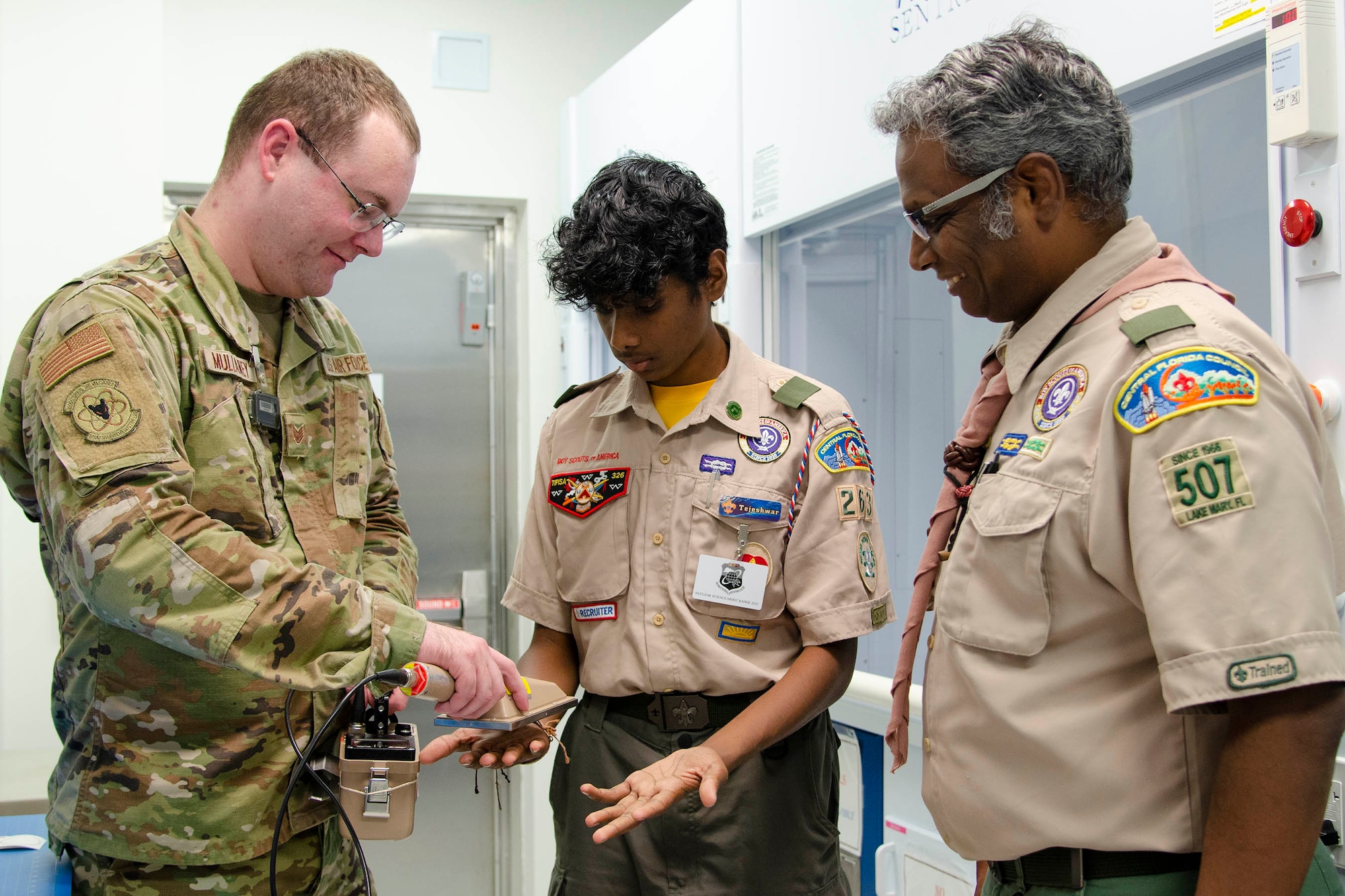 Staff Sgt. John Mullaney II, a scientific applications specialist at the U.S. Air Force Radiochemistry Lab at Patrick Space Force Base, Fla., uses an Alpha/Beta Survey Meter to check for potential contamination on the hands of Tejeshwar Umamaheswar, a Boy Scout from Troop 263 in Lake Mary, Fla., as his father and Scout Master, Umamaheswar Kasinathan, looks on. Teja was one of 62 scouts who traveled to the Air Force Technical Applications Center Oct. 22, 2022 to earn the Nuclear Science Merit Badge with help from Airmen who work at the Department of Defense’s sole nuclear treaty monitoring center. (U.S. Air Force photo by Susan A. Romano)