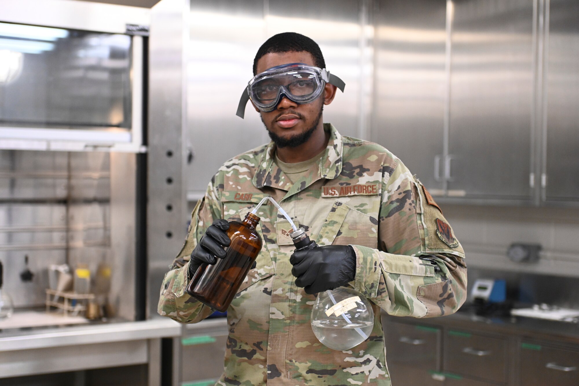 A photo of an Airman holding two instruments used in a fuel lap, wearing goggles.